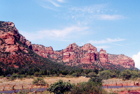 [Cliffs of layered red rock with dark green spots on them spans the entire image. There is more vegetation at the lower levels of the cliffs. Photo was taken at the floor level of the cliffs looking at them from a distance.]